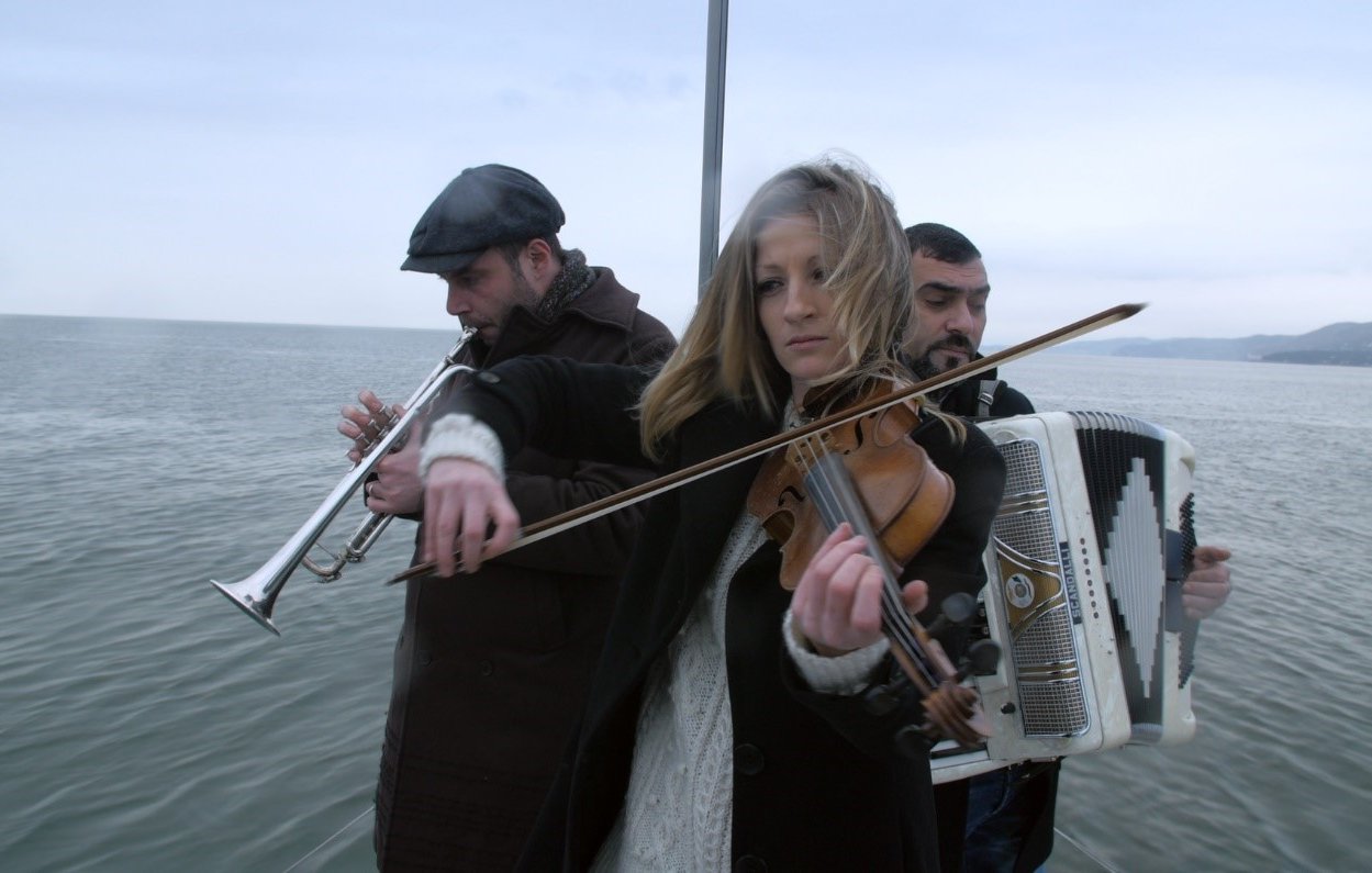 L'immagine mostra tre musicisti che suonano su una piattaforma sopra l'acqua. Una giovane donna al centro suona il violino, mentre indossa un maglione chiaro e un cappotto scuro. A sinistra c'è un uomo che suona un flauto ed è vestito con un cappello e un cappotto scuro. A destra, un altro uomo suona l'accordeon. Lo sfondo è caratterizzato da acque calme e un cielo nuvoloso, creando un'atmosfera suggestiva e malinconica.