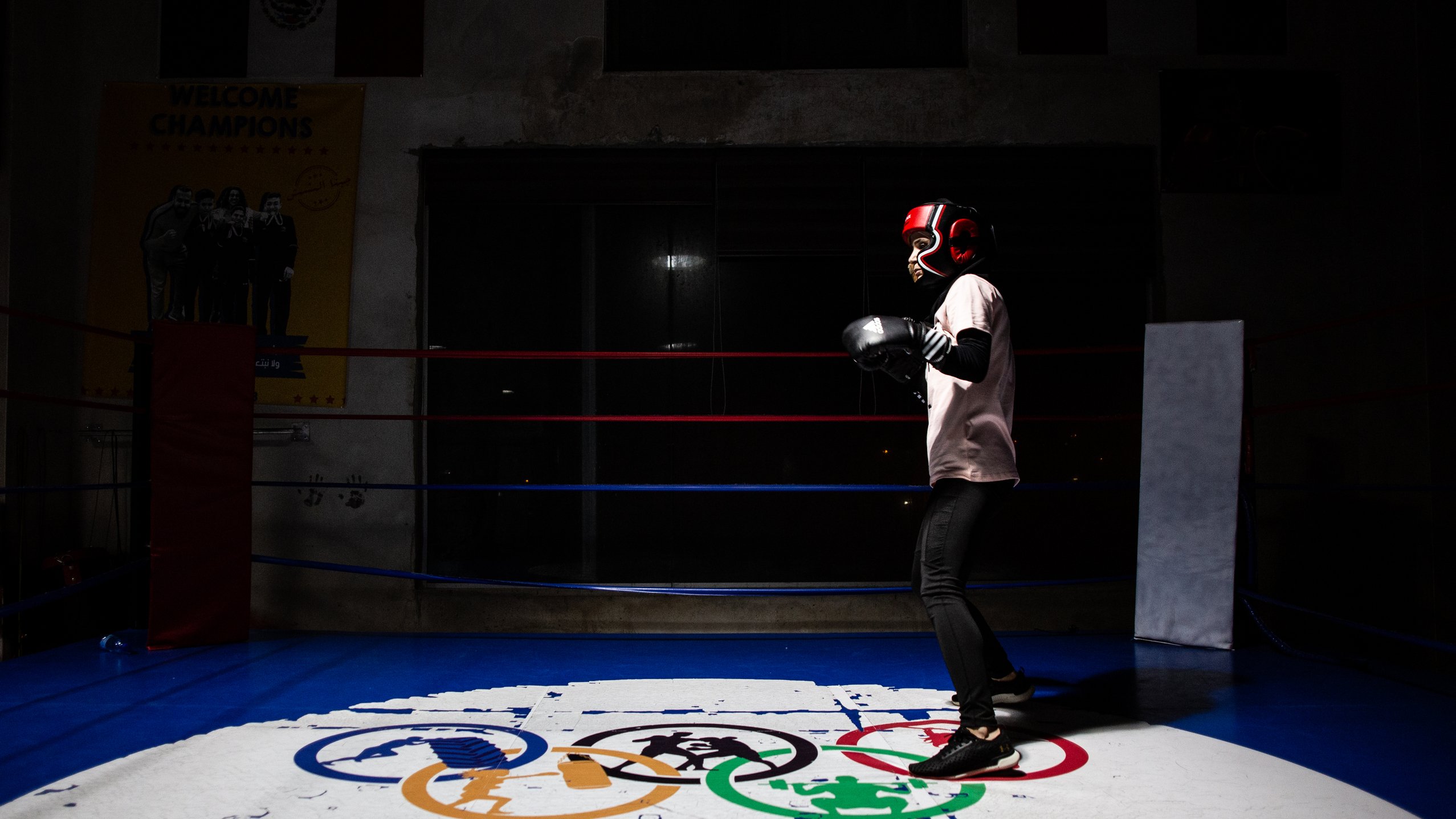L'immagine mostra un giovane pugile che indossa i guantoni e un casco protettivo, mentre si allena in un ring da boxe. Lo sfondo è parzialmente illuminato, mettendo in risalto il pugile in azione. Sotto di lui, si trova un logo con i simboli olimpici e la scritta "ELBARRIO", che suggerisce un ambiente dedicato allo sport e alla formazione. La scena ha un'atmosfera di concentrazione e determinazione.