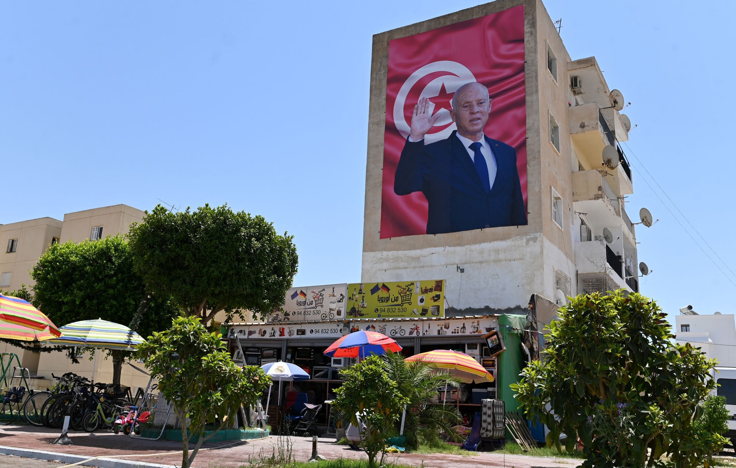 L'immagine mostra un grande murales su un edificio, raffigurante una figura maschile che saluta con una mano alzata. Sullo sfondo, è presente una bandiera che presenta i colori e il simbolo della Tunisia. Nella parte inferiore si possono vedere alcuni ombrelloni colorati, accanto a un'area con biciclette e persone, il tutto immerso in un ambiente urbano. Il cielo è sereno e luminoso, suggerendo una giornata di sole.