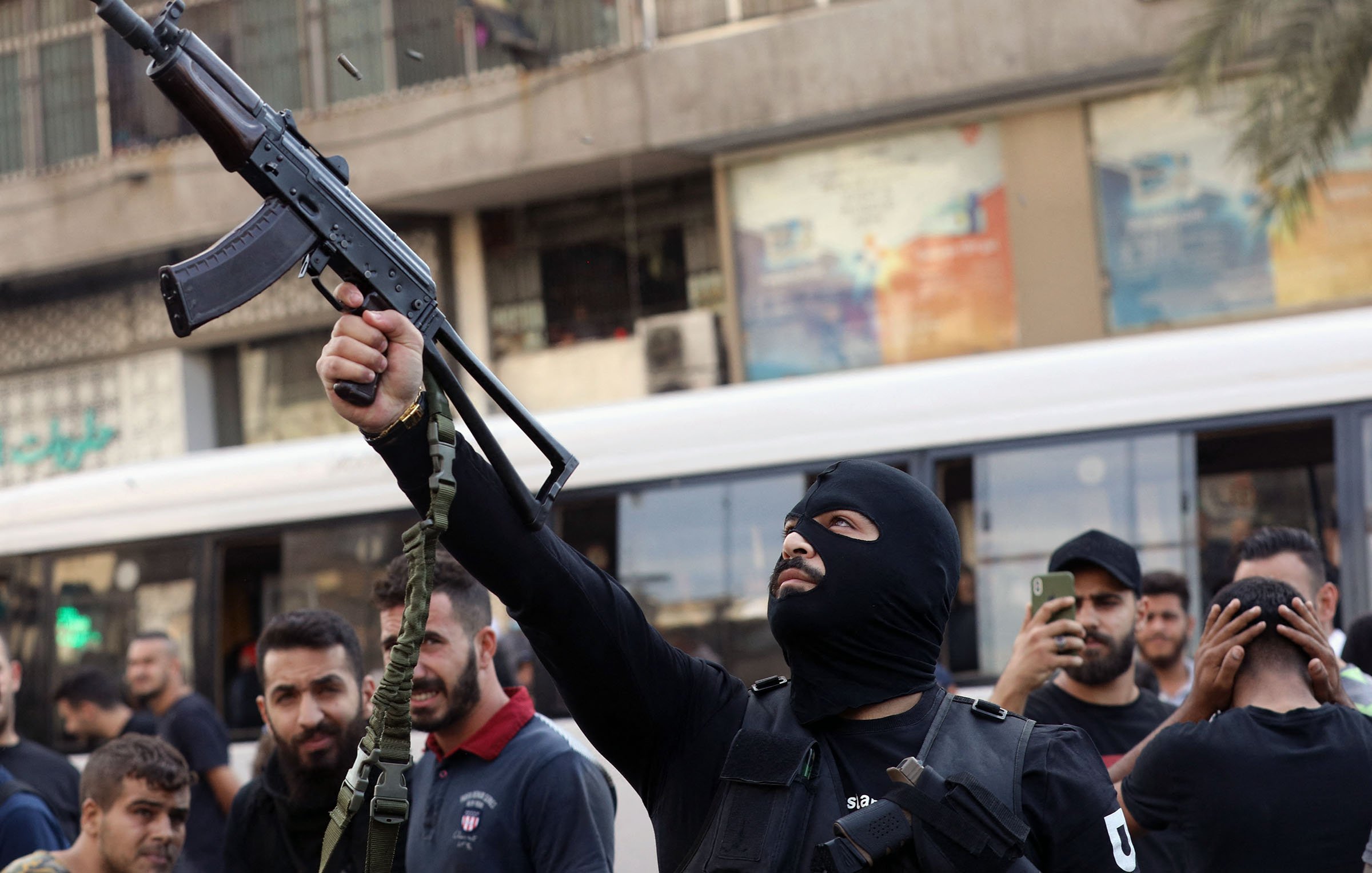 L'immagine mostra un uomo mascherato che brandisce un fucile automatico in una strada affollata. Si possono vedere altre persone intorno a lui, alcune delle quali sembrano sostenere o osservare l'azione. L'ambiente circostante è urbanizzato, con edifici e un autobus che appare in lontananza. L'atmosfera sembra carica di tensione, suggerendo un contesto di protesta o conflitto.