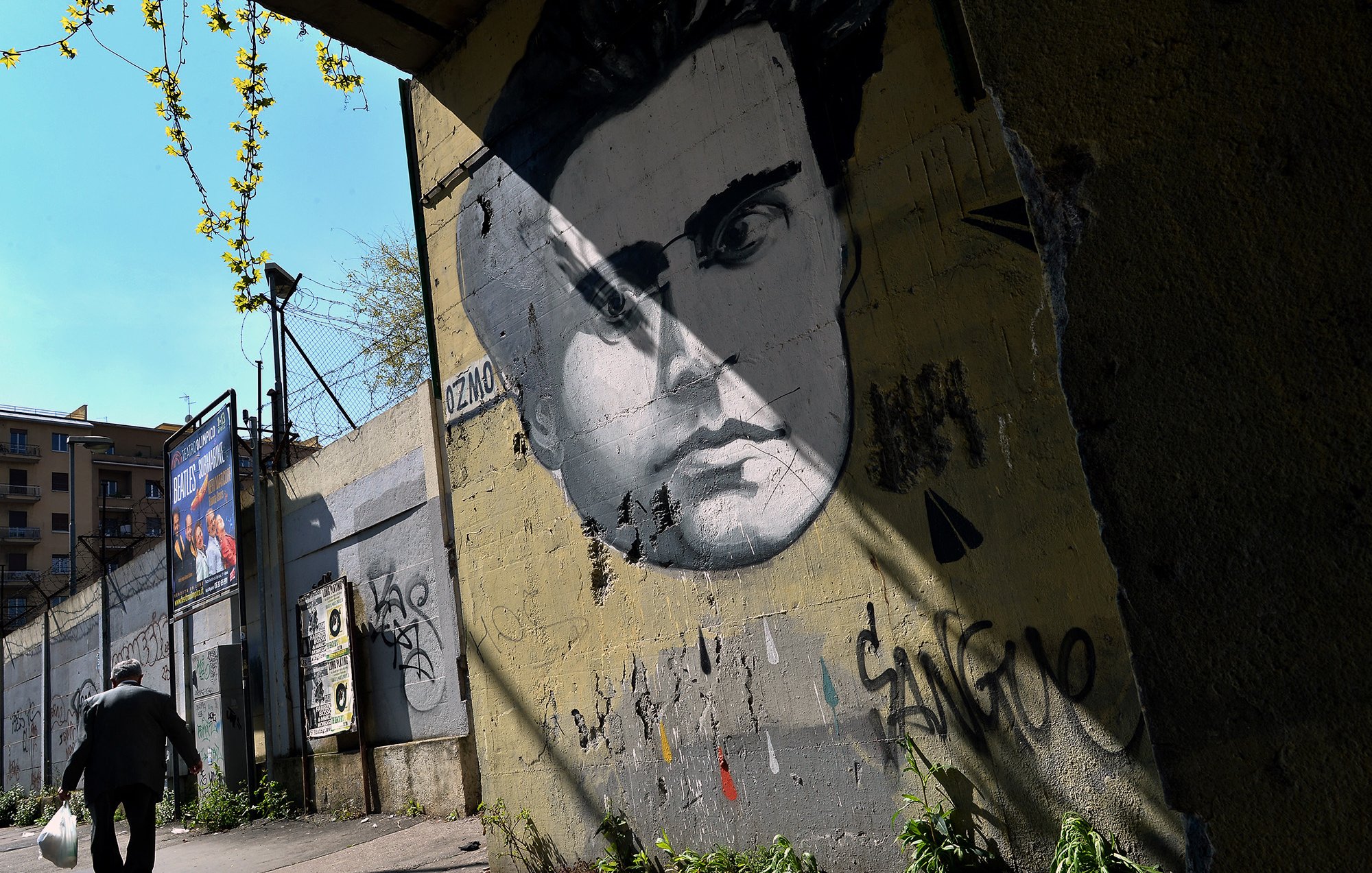 L'immagine mostra un murale di un volto umano, molto probabilmente di un uomo, dipinto su un muro grigio. Il viso ha lineamenti distintivi e intensi, con un'espressione che attira l'attenzione. Sullo sfondo, si può vedere una strada o un marciapiede con un uomo che cammina, mentre alcuni elementi di verde e graffiti decorano la scena. La luce del sole crea una serie di ombre, accentuando l'atmosfera urbana del luogo.