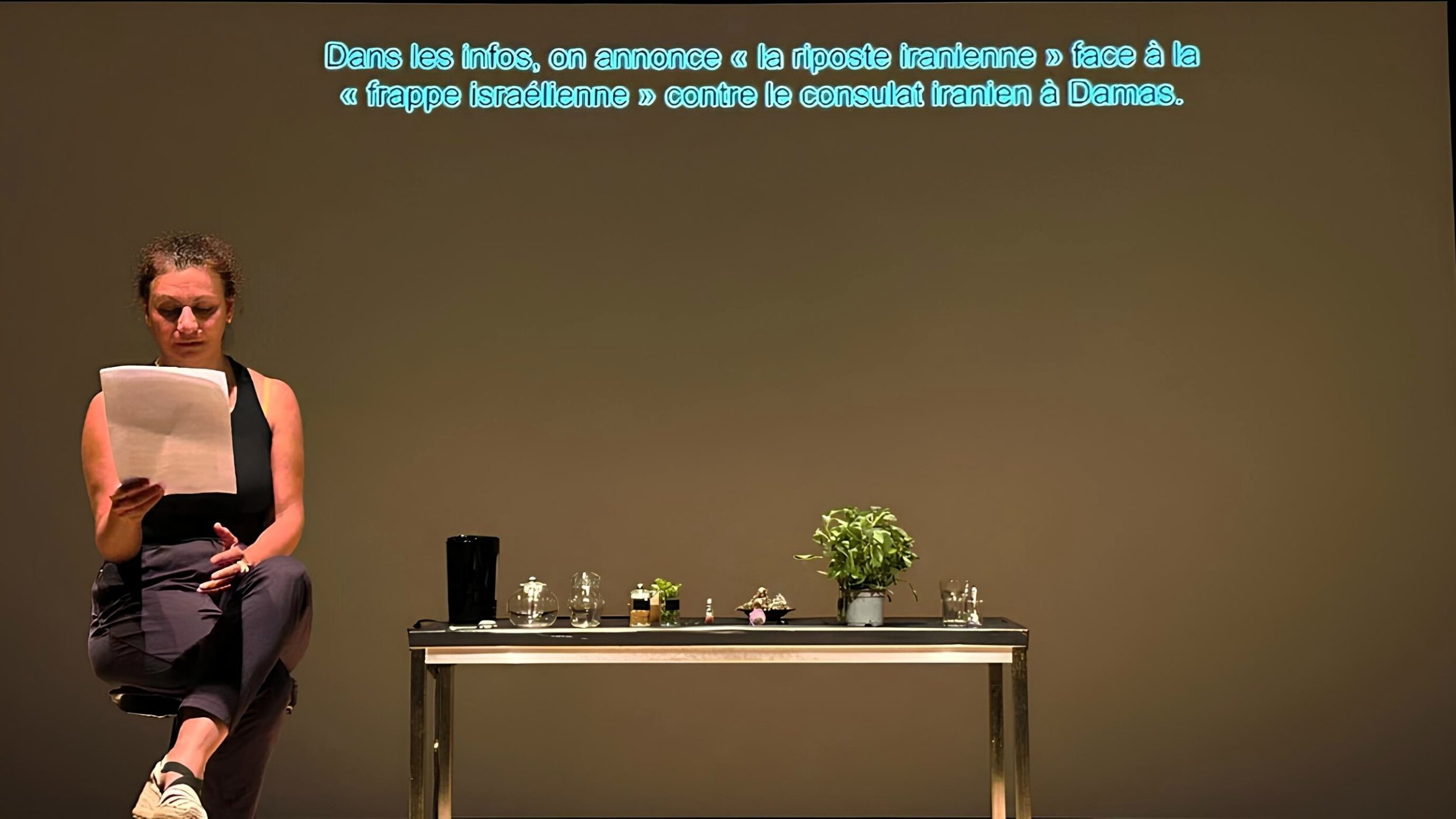 Una donna legge appunti davanti a un tavolo con piante e oggetti, su uno sfondo neutro.