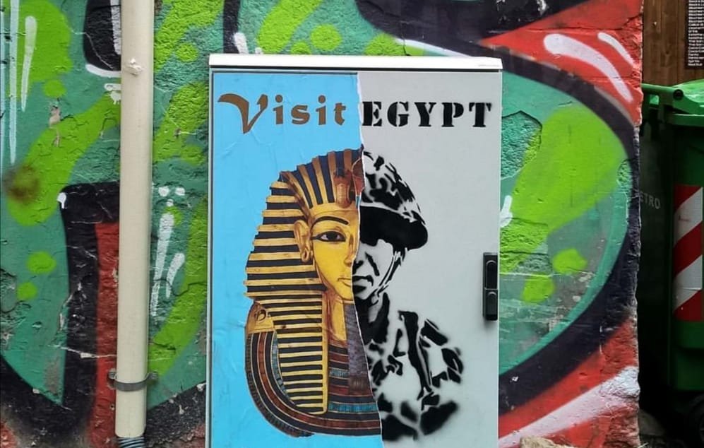 L'immagine mostra un armadietto metallico dipinto con un murale che presenta due volti distinti. A sinistra, c'è un'immagine stilizzata di un faraone, accompagnata da un invito a "Visit Egypt" e "Enjoy the". A destra, c'è un ritratto che sembra rappresentare un soldato o una figura autoritaria, con la parola "REGIME" sopra di esso. Lo sfondo è colorato e impreziosito da graffiti, creando un contrasto tra i due soggetti. La composizione sembra riflettere una critica sociale o politica legata all'Egitto.