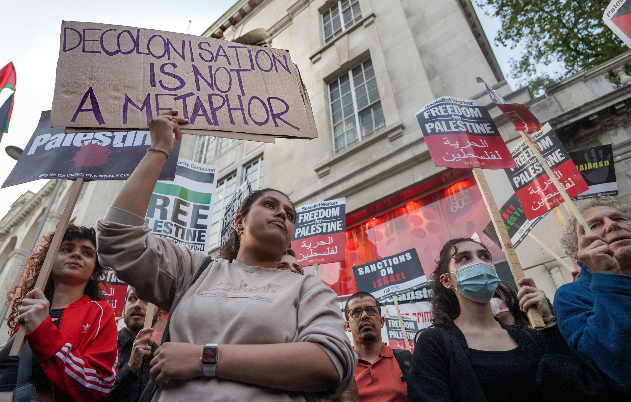 L'immagine mostra un gruppo di persone in una manifestazione, molte delle quali tengono in mano cartelli con frasi di protesta. Una giovane donna al centro tiene un cartello che dice "La decolonizzazione non è una metafora". Sullo sfondo, ci sono altri cartelli con scritte come "Libertà per la Palestina" e "Sanzioni a Israele". I partecipanti sembrano esprimere il loro sostegno per la causa palestinese, mentre l'atmosfera è di attivismo e impegno sociale.