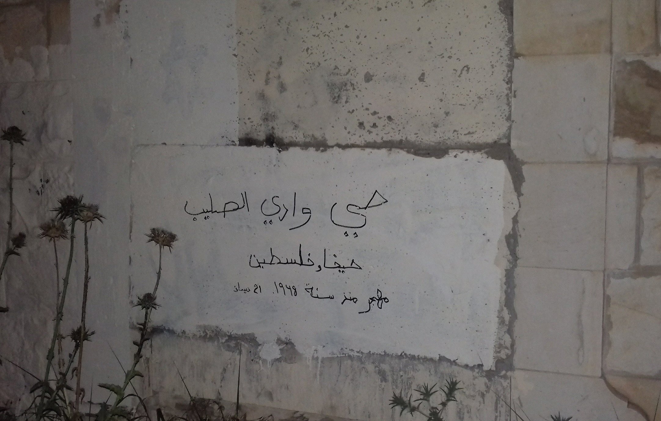 L'immagine mostra una parete di pietra con un'iscrizione in arabo. La scritta fa riferimento a un luogo specifico, con delle date evidenti, probabilmente legate alla storia palestinese. Attorno alla scritta si notano delle erbacce, che suggeriscono che il posto non sia in uso o mantenga un aspetto trascurato. La scena evoca un senso di abbandono e memoria.