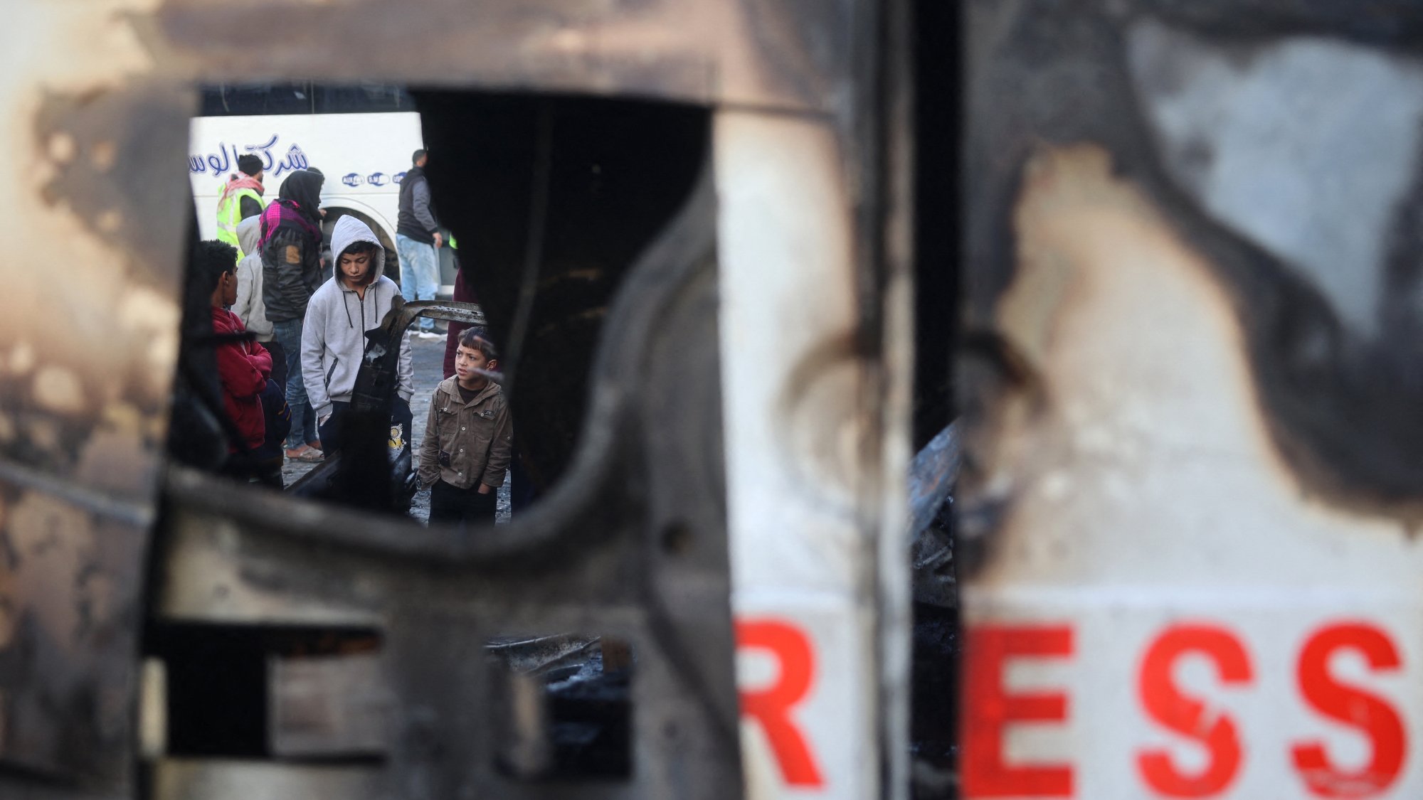 L'immagine mostra una scena cupa e difficile, probabilmente in un contesto di conflitto o crisi. Si vede un gruppo di persone che si trovano in un'area devastata, ricevendo assistenza o cercando di mettersi in salvo. Parte della scena è incorniciata da un oggetto danneggiato, che contribuisce a esprimere la tensione e la difficoltà della situazione. Le persone nel gruppo appaiono preoccupate e potrebbero essere in attesa di aiuto o di informazioni.