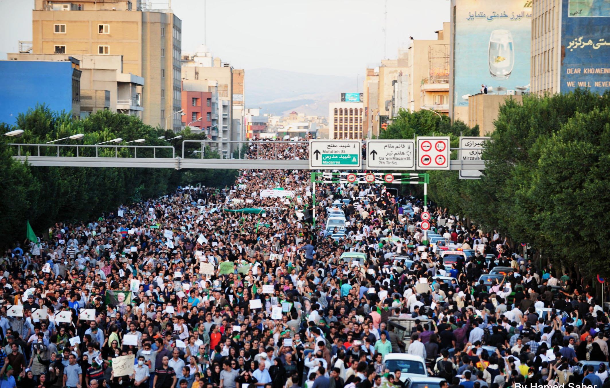 L'immagine mostra una grande folla di manifestanti in una strada cittadina. Le persone sembrano riunite per un evento significativo, con molti che portano cartelli e striscioni. I palazzi al bordo della strada sono visibili, così come alcune automobili parcheggiate. La scena trasmette una forte sensazione di unione e mobilitazione collettiva. Le strade sono affollate e l'atmosfera è intensa, suggerendo che si tratta di una protesta o di una manifestazione pubblica.