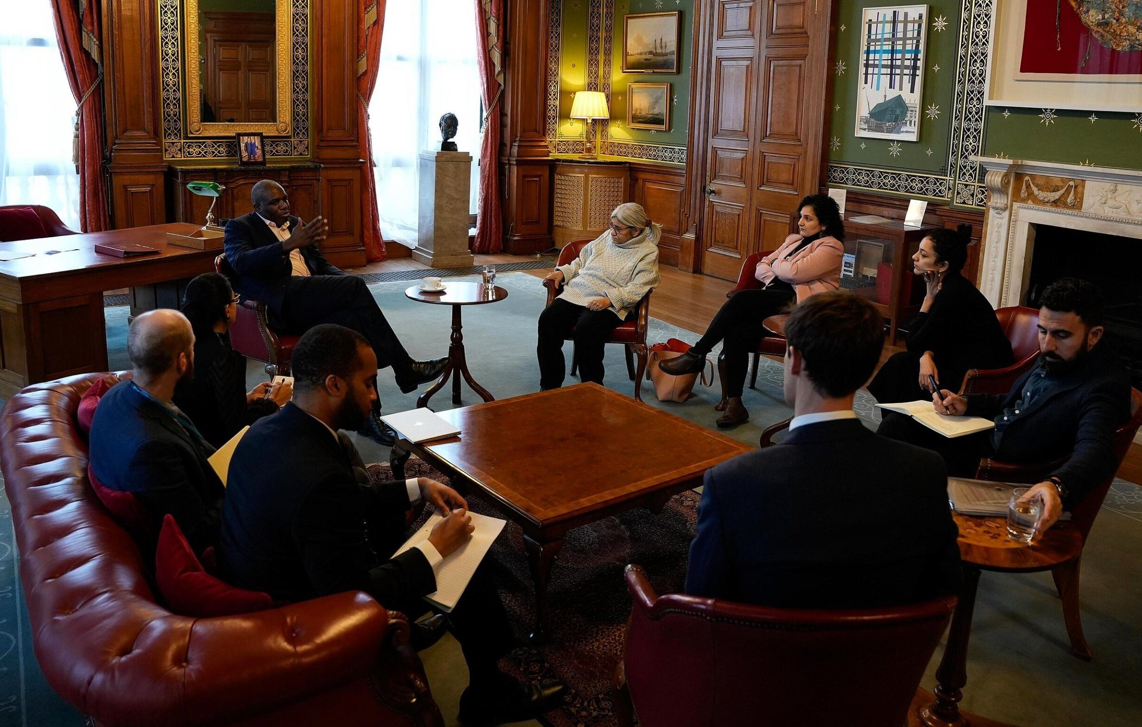 Nell'immagine si vede un incontro formale all'interno di una stanza ben arredata, con pareti in legno e decorazioni artistiche. Un gruppo di persone è riunito attorno a un tavolino centrale. Alcuni partecipanti sono seduti su poltrone, mentre altri sono seduti su uno sgabello o in posizioni più informali. Alcuni hanno in mano blocchi note o penne, suggerendo che stanno prendendo appunti. La luce naturale entra attraverso le finestre, creando un'atmosfera accogliente. Sullo sfondo, si intravedono alcune opere d'arte e un camino, che aggiungono un tocco elegante all'ambiente.