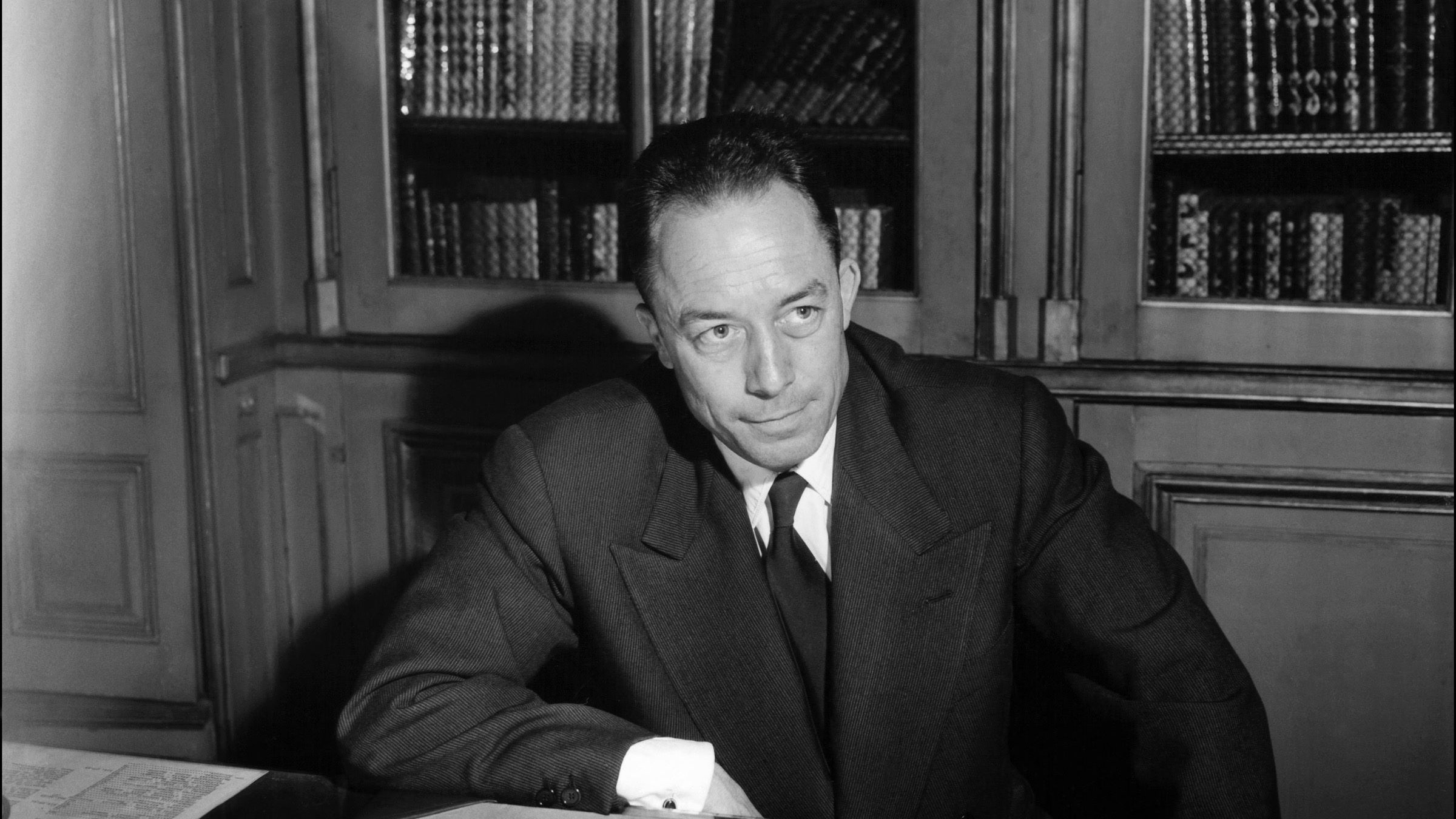 L'immagine mostra un uomo seduto a un tavolo, con dei documenti sparsi davanti a lui. Indossa un abito scuro con una cravatta, e appare pensieroso mentre guarda verso un punto oltre la fotocamera. Sullo sfondo si intravedono alcune librerie con libri. L'illuminazione è elegante, creando un'atmosfera seria e riflessiva.