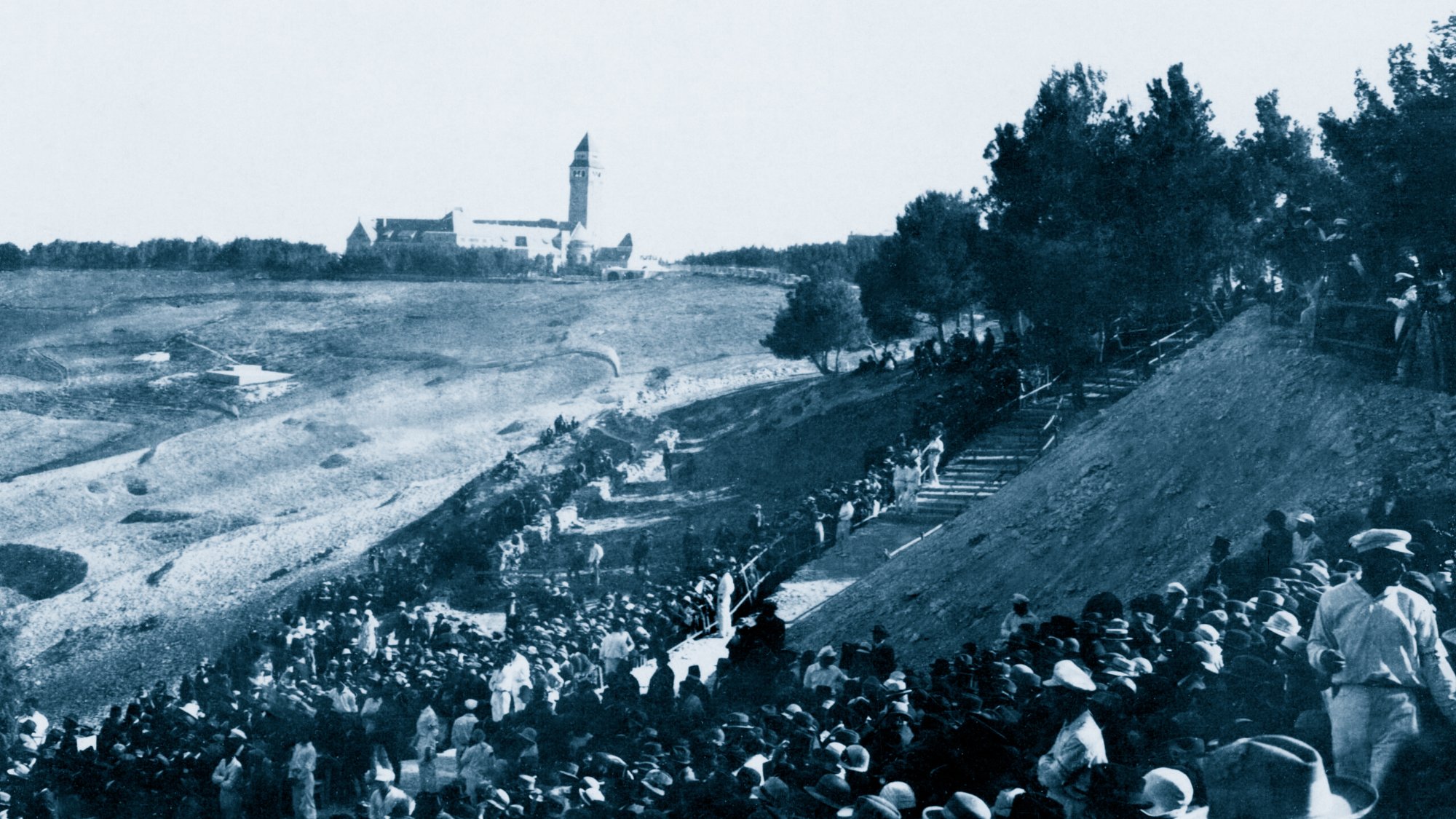 Un grande raduno di persone all'aperto, con un edifico distante e alberi circostanti.