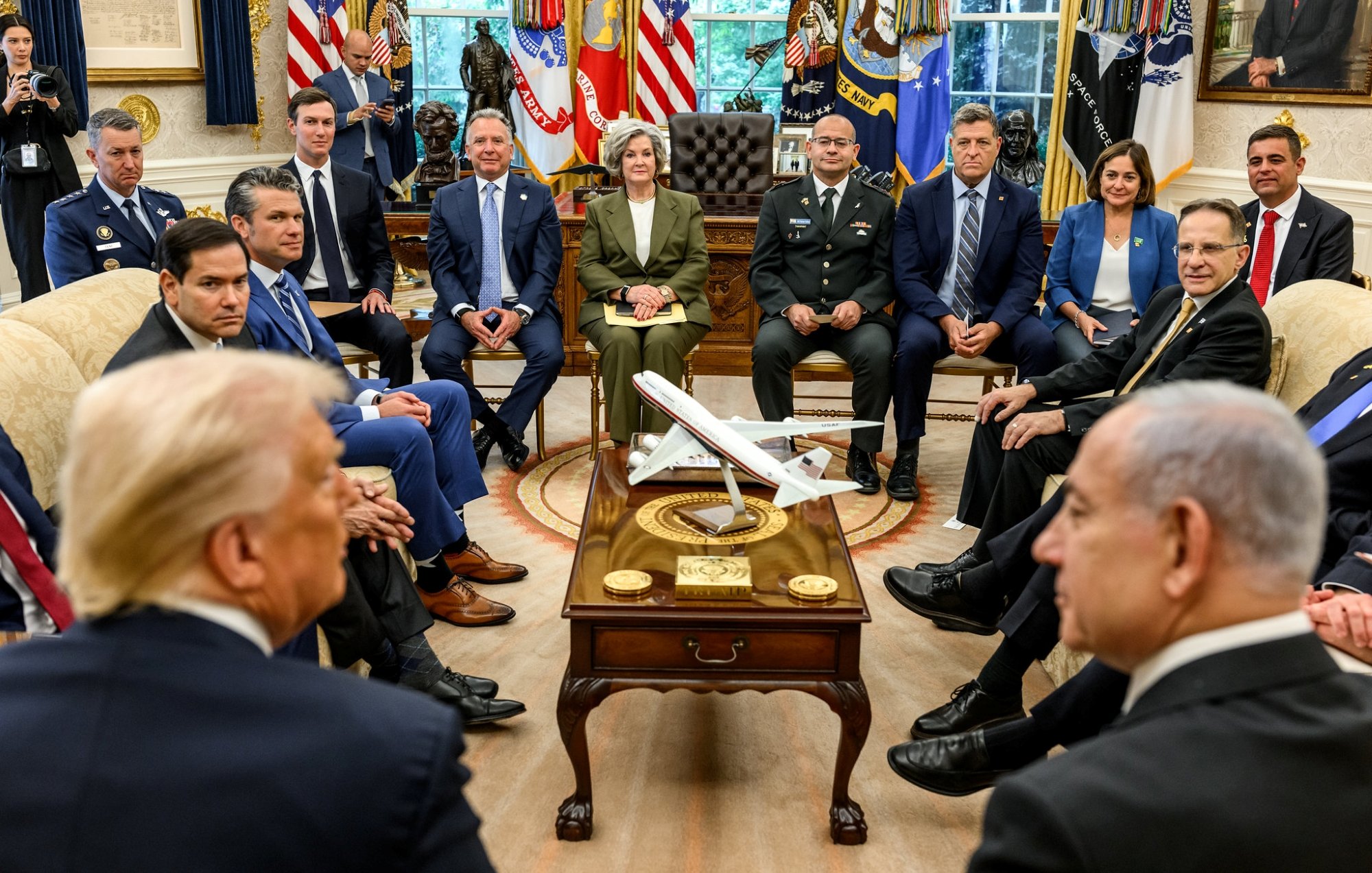 In una sala con bandiere, vari uomini e donne seduti attorno a un tavolo con un aereo in miniatura.