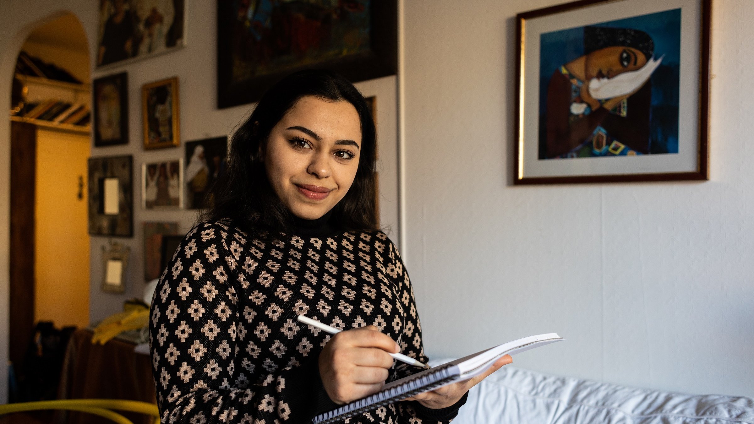 Nell'immagine, una giovane donna sorride mentre tiene un taccuino e una penna in mano. Indossa un maglione con un motivo geometrico. Sullo sfondo si può vedere una stanza con pareti decorate da vari quadri e fotografie. C'è un divano bianco e una sedia gialla, che aggiungono un tocco di colore all'ambiente. L'atmosfera è calda e accogliente.