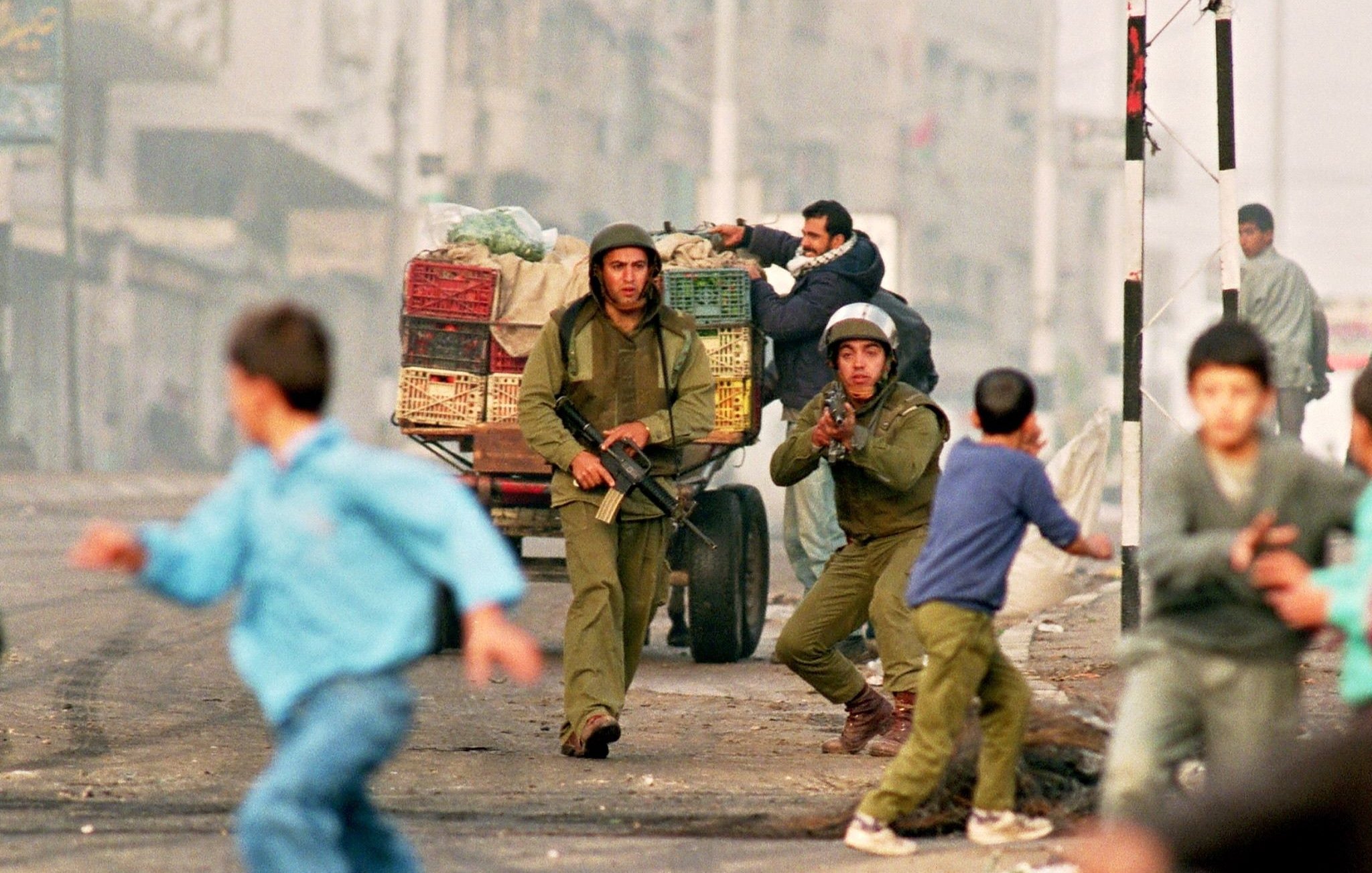 L'immagine ritrae una scena di conflitto in un'area urbana. Due soldati in uniforme stanno correndo, uno dei quali sembra puntare un'arma. Dietro di loro, un carro con casse di frutta è visibile. In primo piano, dei bambini stanno fuggendo, mostrando un'espressione di paura. L'ambiente circostante appare caotico, con una luce che suggerisce un'atmosfera di tensione. La scena comunica chiaramente una situazione di crisi e instabilità.