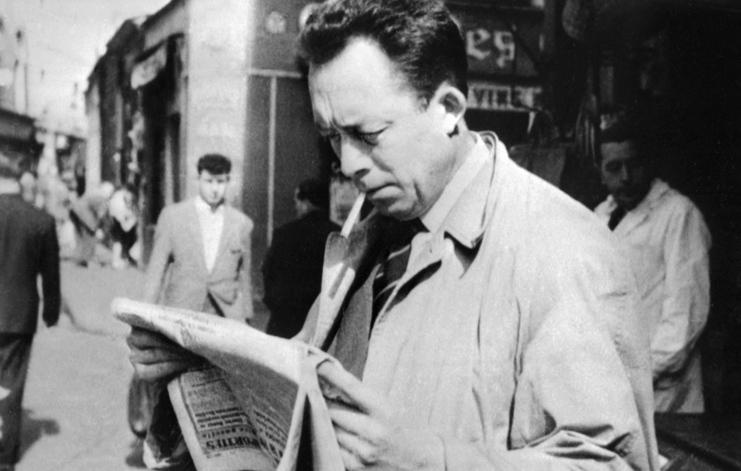 L'immagine ritrae un uomo in primo piano che legge un giornale, con una sigaretta tra le labbra. Indossa un soprabito chiaro e sembra assorto nella lettura. Sullo sfondo, si possono vedere altre persone che camminano per strada, in un contesto urbano. L'atmosfera è retro e richiama un'epoca passata, con un'illuminazione naturale e dettagli architettonici tipici di quel periodo.