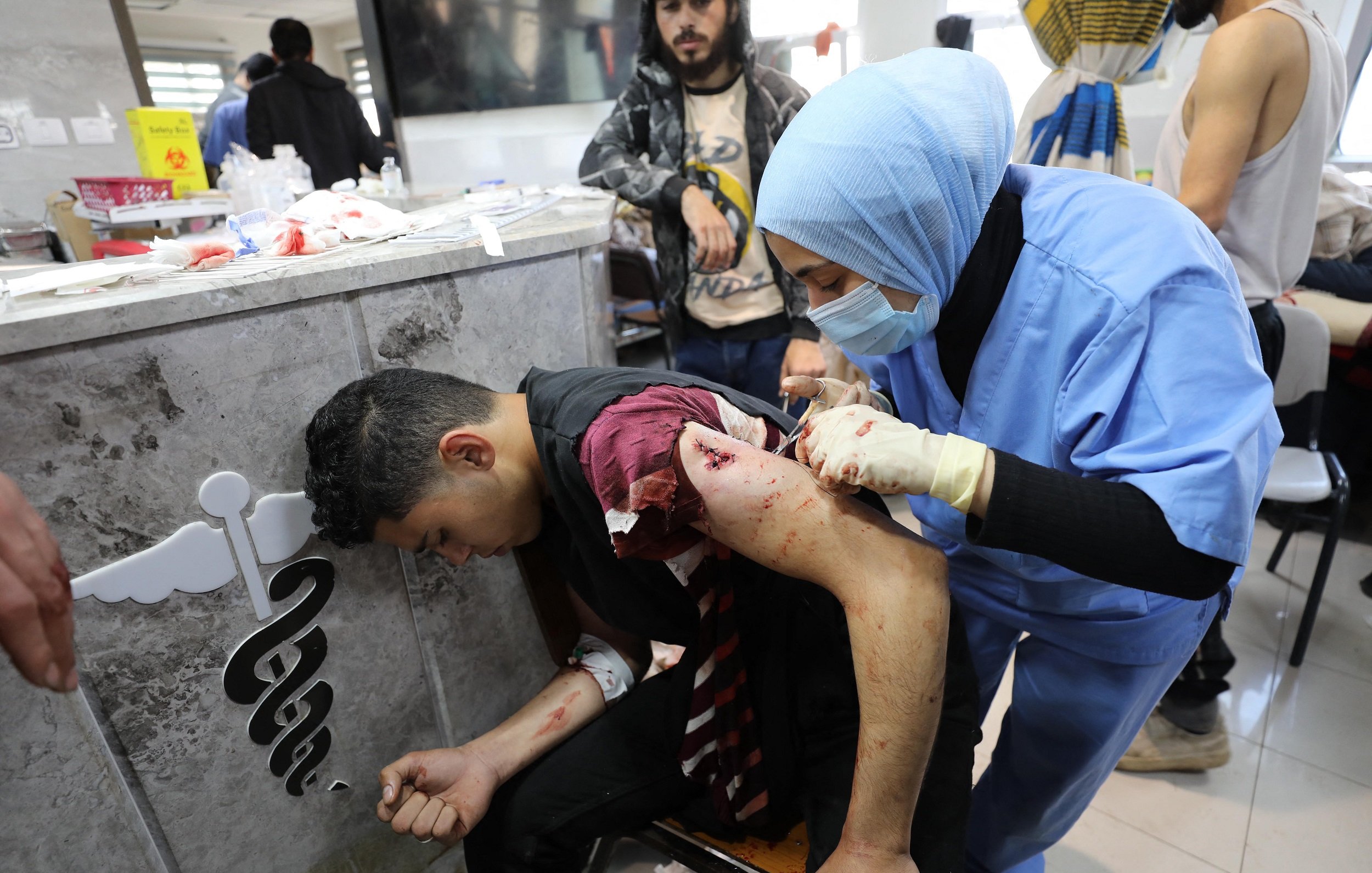 L'immagine mostra una scena in un ambiente sanitario. Un giovane uomo, visibilmente ferito, è seduto su una sedia mentre una professionista sanitaria, vestita con una divisa blu e con guanti, sta medicando una sua ferita sul braccio. Il suo volto mostra preoccupazione, e l'atmosfera appare tesa. Sullo sfondo si possono vedere altre persone e attrezzature mediche, suggerendo un contesto di emergenza.