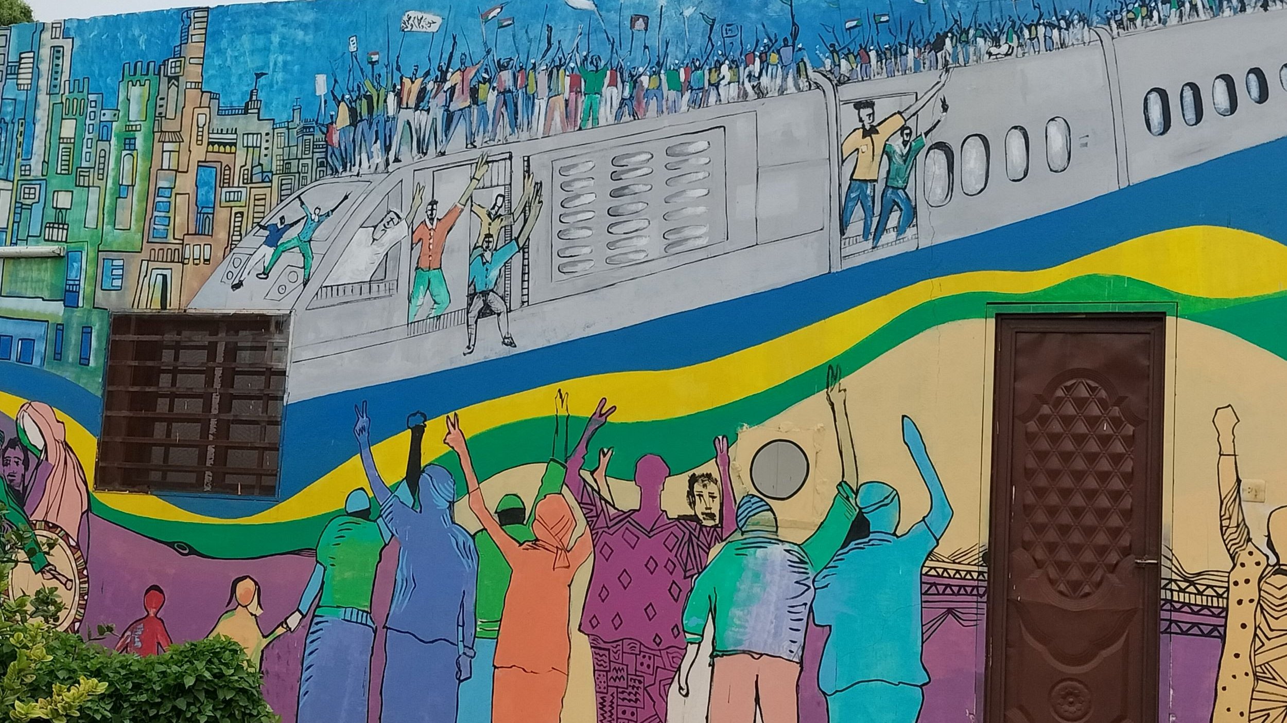 L'immagine rappresenta un murale colorato su un muro. La scena ritrae una folla di persone che sembra celebrare o protestare, con alcune figure che sollevano le braccia in segno di gioia o di richiesta. Sullo sfondo, c'è un aereo che si staglia sopra una città stilizzata, caratterizzata da edifici e una vivace palette di colori. I colori utilizzati nel murale sono brillanti e vivaci, riflettendo un senso di energia e movimento. La composizione comunica un forte messaggio di unità e di movimento collettivo.