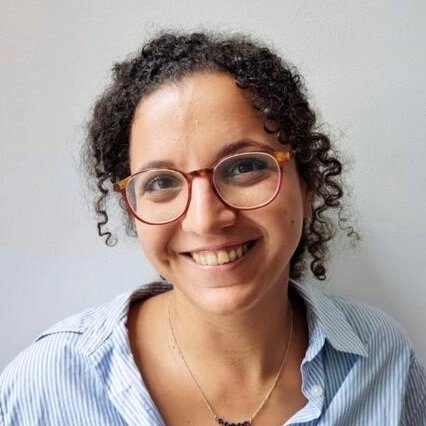 L'image montre une femme souriante avec des cheveux bouclés et des lunettes. Elle porte une chemise à rayures et un collier. L'arrière-plan est simple et clair, mettant en valeur son expression joyeuse.