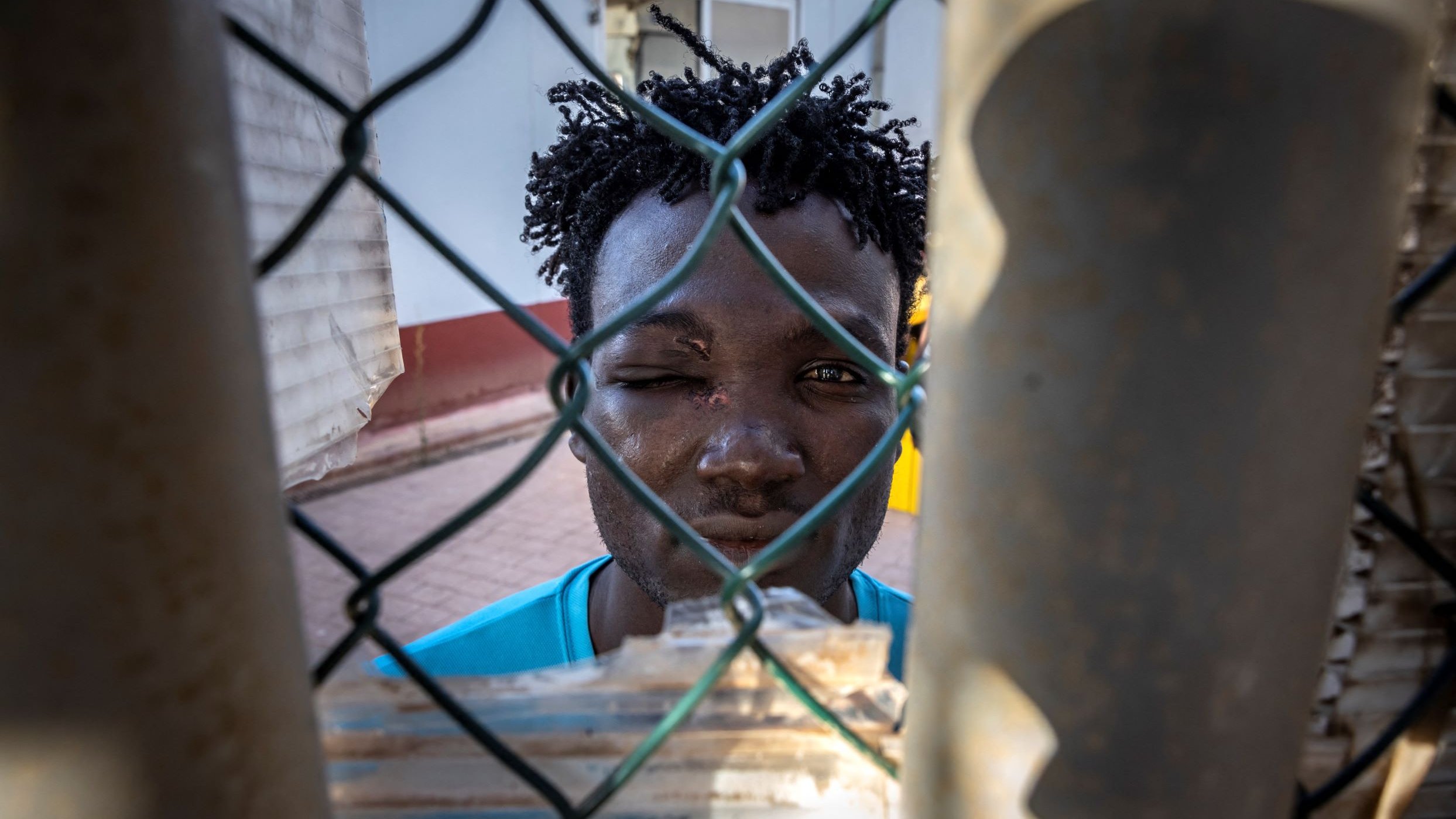 L'immagine mostra un giovane uomo che si affaccia attraverso una rete di recinzione. Ha i capelli ricci e un'espressione seria. Il suo viso è parzialmente coperto da una rete metallica, il che crea un effetto di separazione. Sullo sfondo, si intravede un'architettura semplice. La luce colpisce il suo volto, mettendo in evidenza i dettagli della sua espressione.