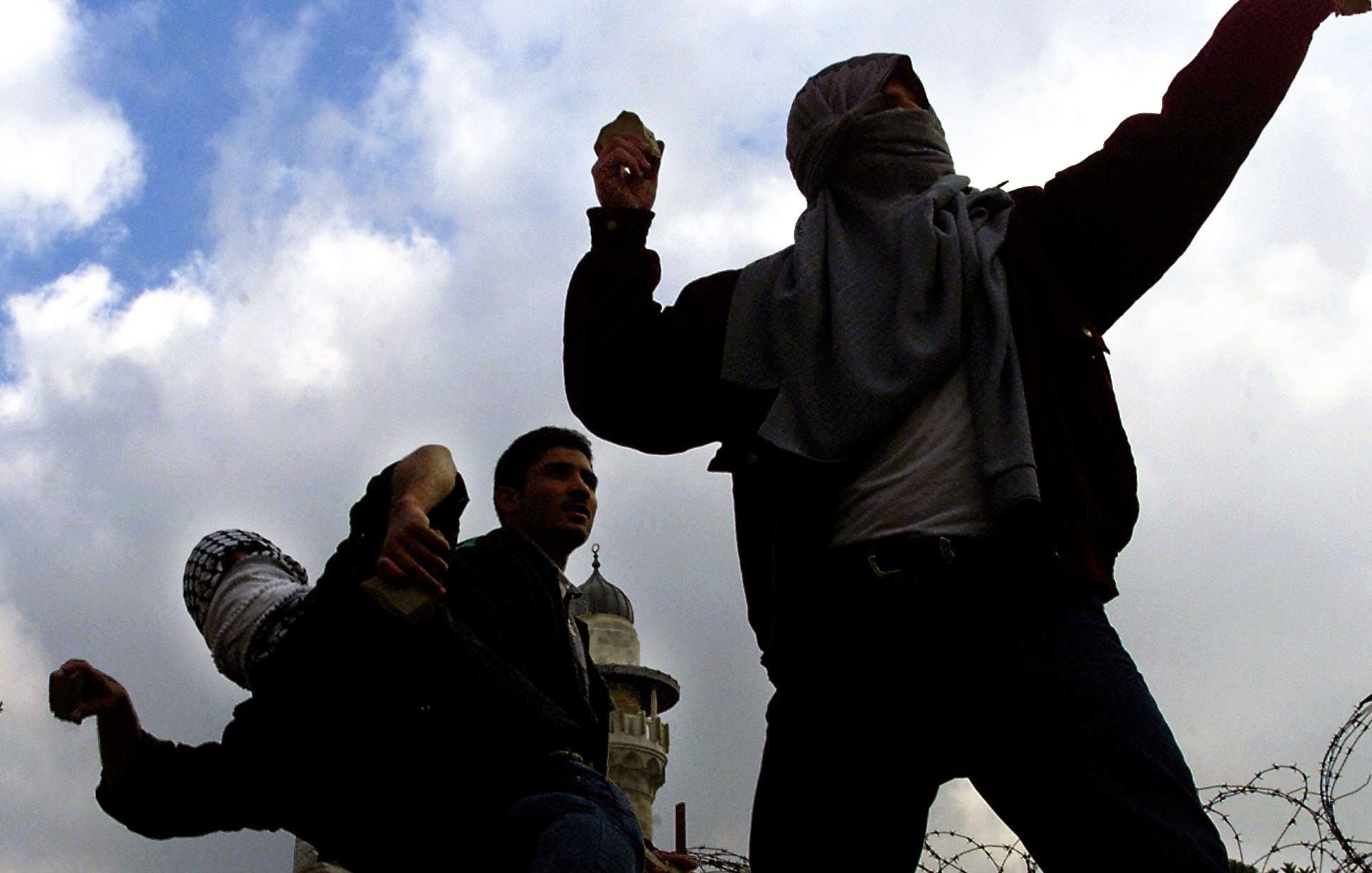 L'immagine mostra un gruppo di persone in una situazione di tensione, probabilmente in un contesto di protesta o conflitto. Alcuni individui sono coperti da sciarpe o fazzoletti, mentre sembrano lanciare dei proiettili, forse pietre. Sullo sfondo si possono vedere strutture architettoniche e un cielo nuvoloso, che contribuiscono all'atmosfera drammatica della scena. La presenza di filo spinato suggerisce una situazione di conflitto o di controllo militare.