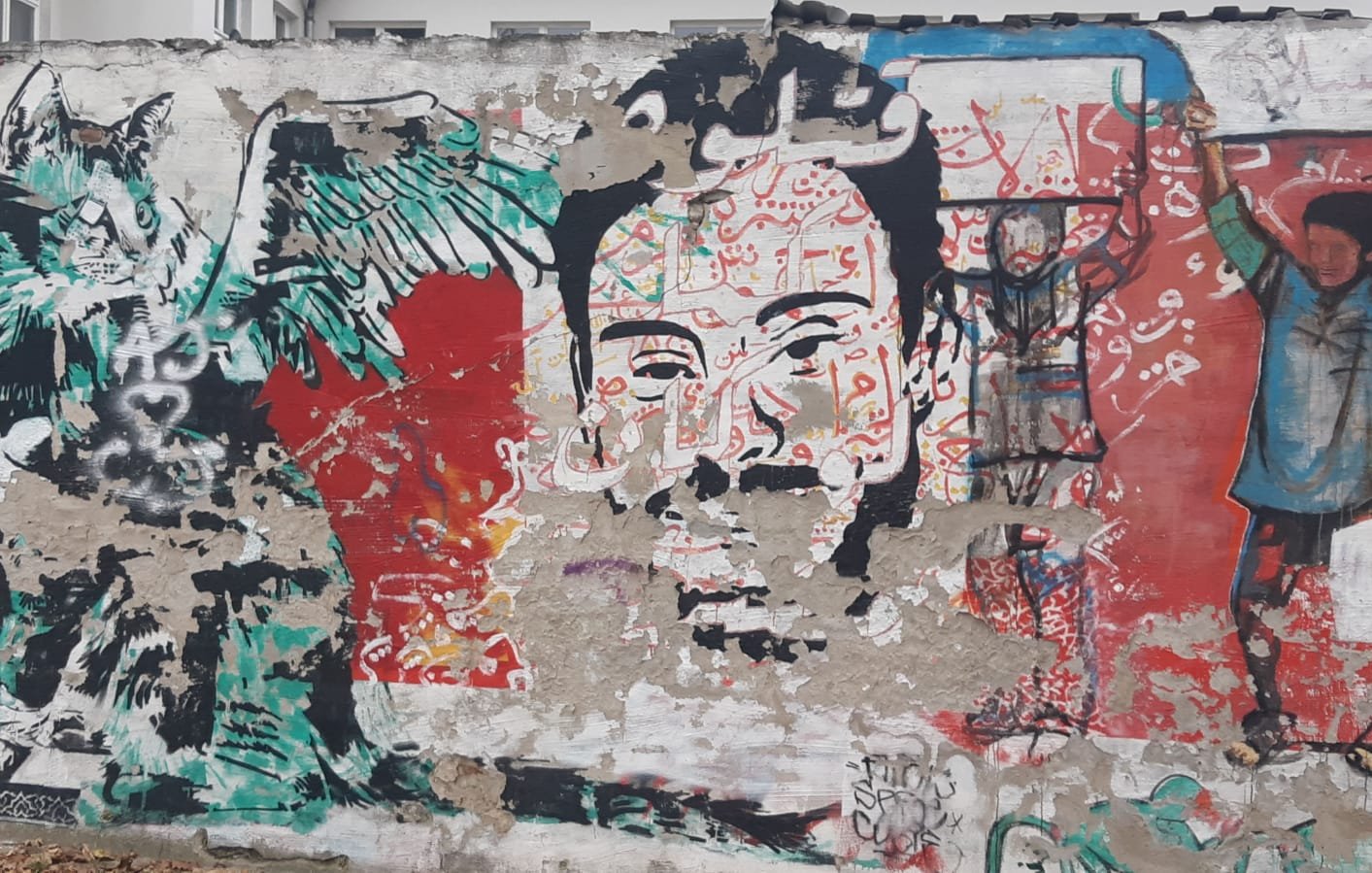 L'immagine mostra un murale su un muro. A sinistra, si può vedere un disegno di una creatura con ali e tratti animaleschi, insieme a elementi astratti e colori vivaci. Al centro, c'è un volto umano stilizzato, con dettagli intricati e un'espressione intensa. Sullo sfondo, si nota un altro personaggio con un gesto di sollevamento delle braccia. La composizione è caratterizzata da una ricca varietà di colori e forme, creando un effetto dinamico e coinvolgente.