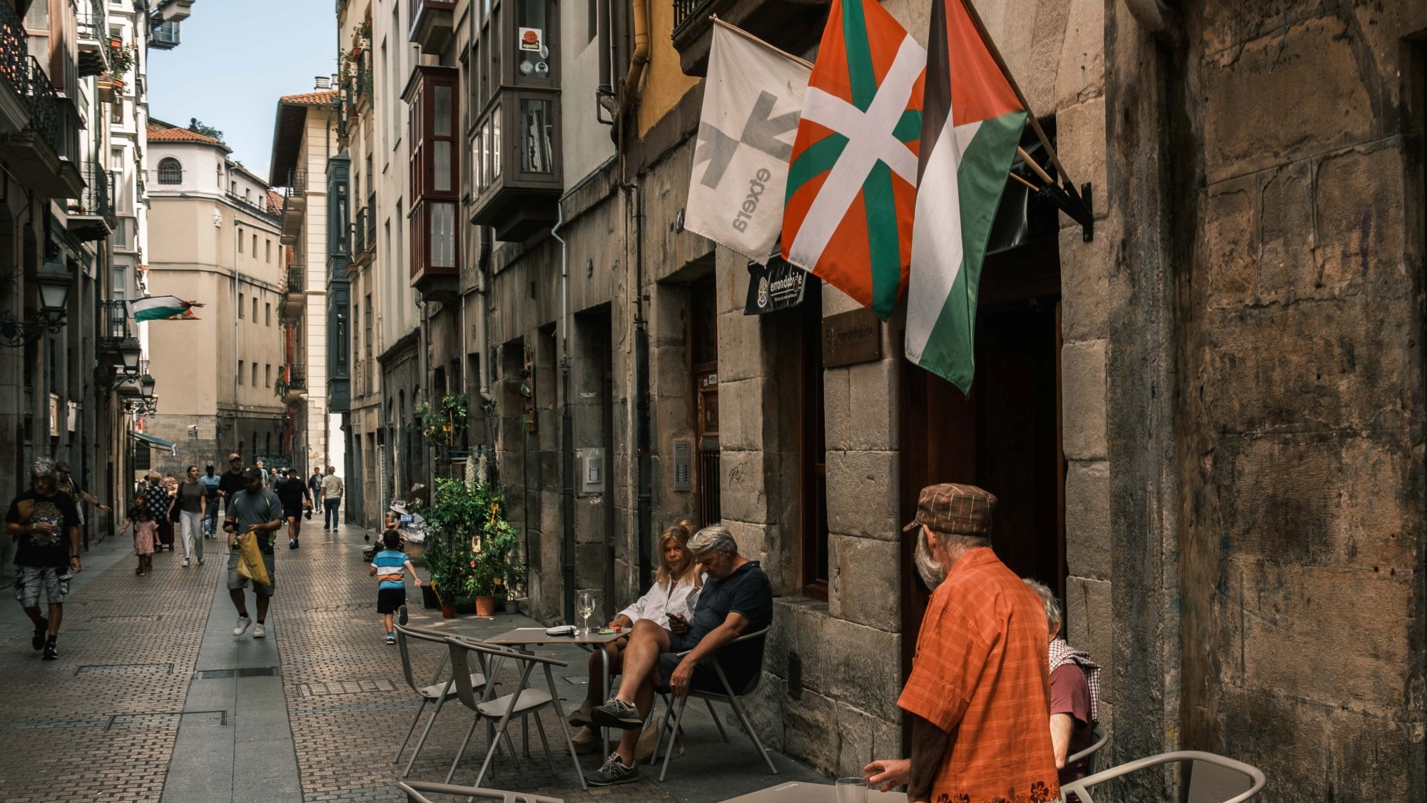 Vicolo affollato con tavolini all'aperto e bandiere che sventolano.