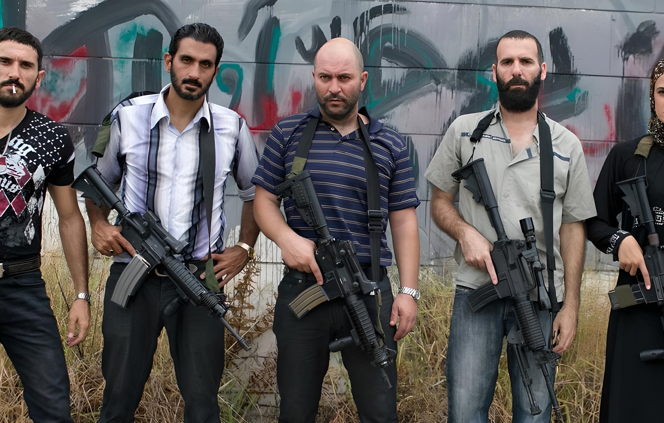 L'immagine mostra un gruppo di cinque persone, tutte in piedi e con armi da fuoco in mano. Quattro di loro hanno un aspetto maschile, mentre una è femminile. Sono vestiti in abiti casual e mostrano espressioni serie. Sullo sfondo si intravedono graffiti, creando un'atmosfera di tensione. L'ambiente sembra essere abbandonato o in degrado, con erba alta che circonda i soggetti.