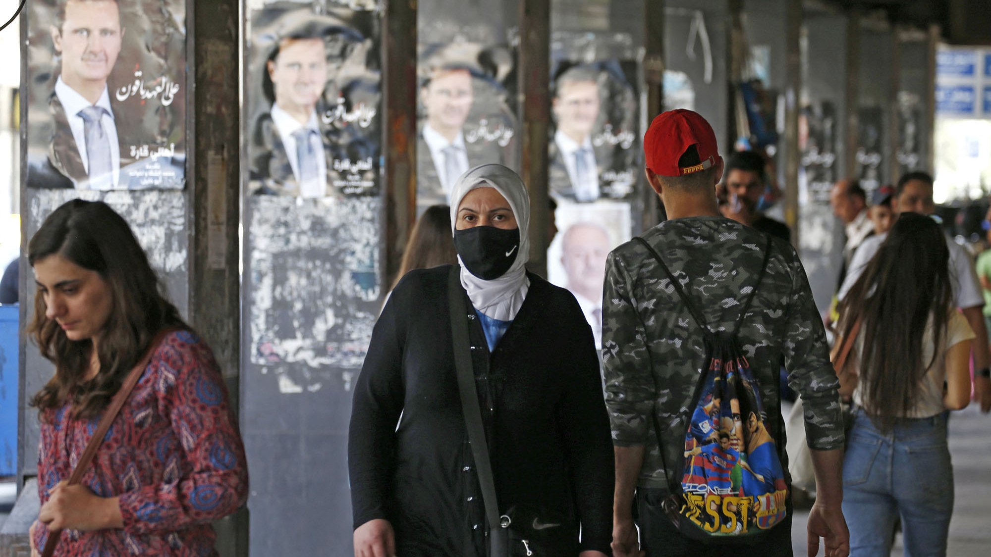 Nell'immagine si vedono diverse persone in un'area urbane, probabilmente un mercato o una strada affollata. Alcuni passanti indossano mascherine, mentre altri non ne hanno. Sullo sfondo, ci sono manifesti con l'immagine di un uomo, presumibilmente un leader politico, appesi alle pareti. L'atmosfera sembra quella di un luogo vivo e multiculturale, con persone di diverse età e stili che si muovono nella zona.