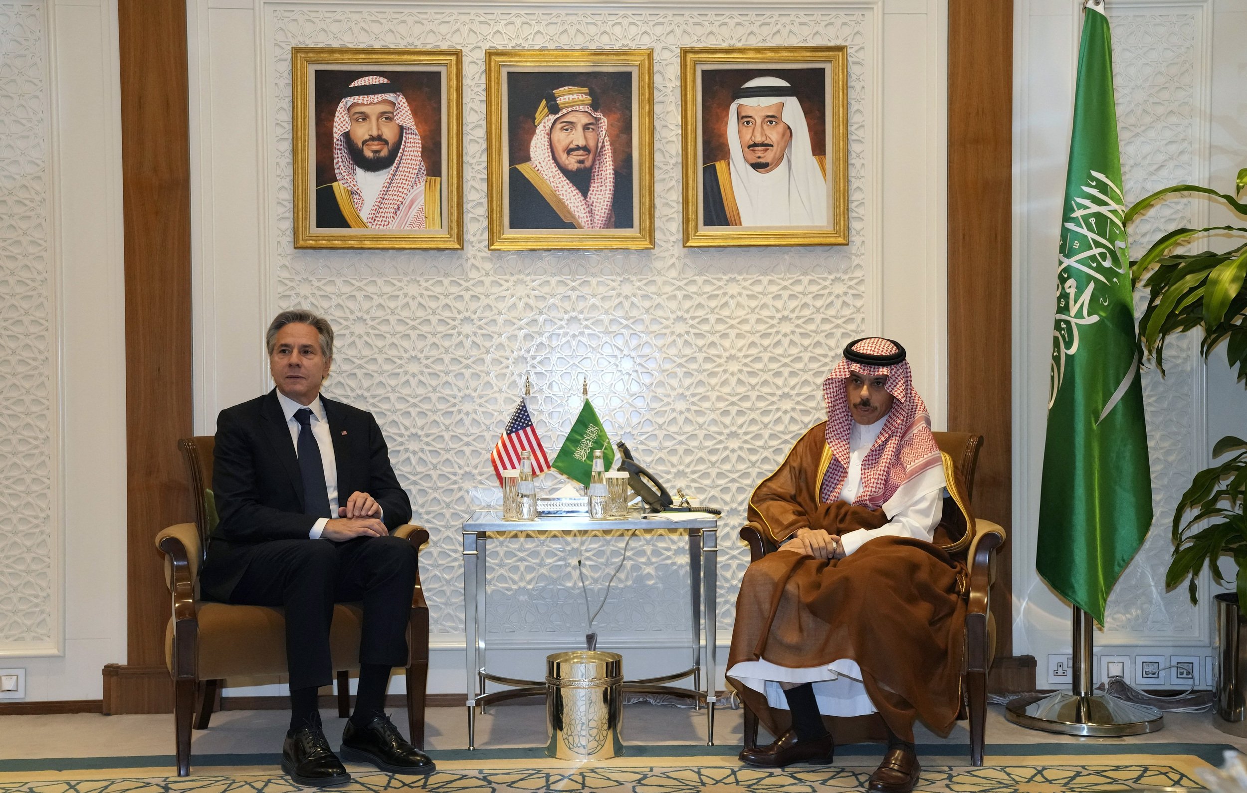 Nell'immagine si possono vedere due uomini seduti uno di fronte all'altro in un ambiente formale. Sullo sfondo ci sono tre ritratti incorniciati, probabilmente di leader sauditi. A sinistra, è presente una bandiera americana e a destra una bandiera saudita. Il tavolo tra i due uomini contiene alcuni oggetti decorativi. L'atmosfera apparente è di dialogo politico o diplomatico.