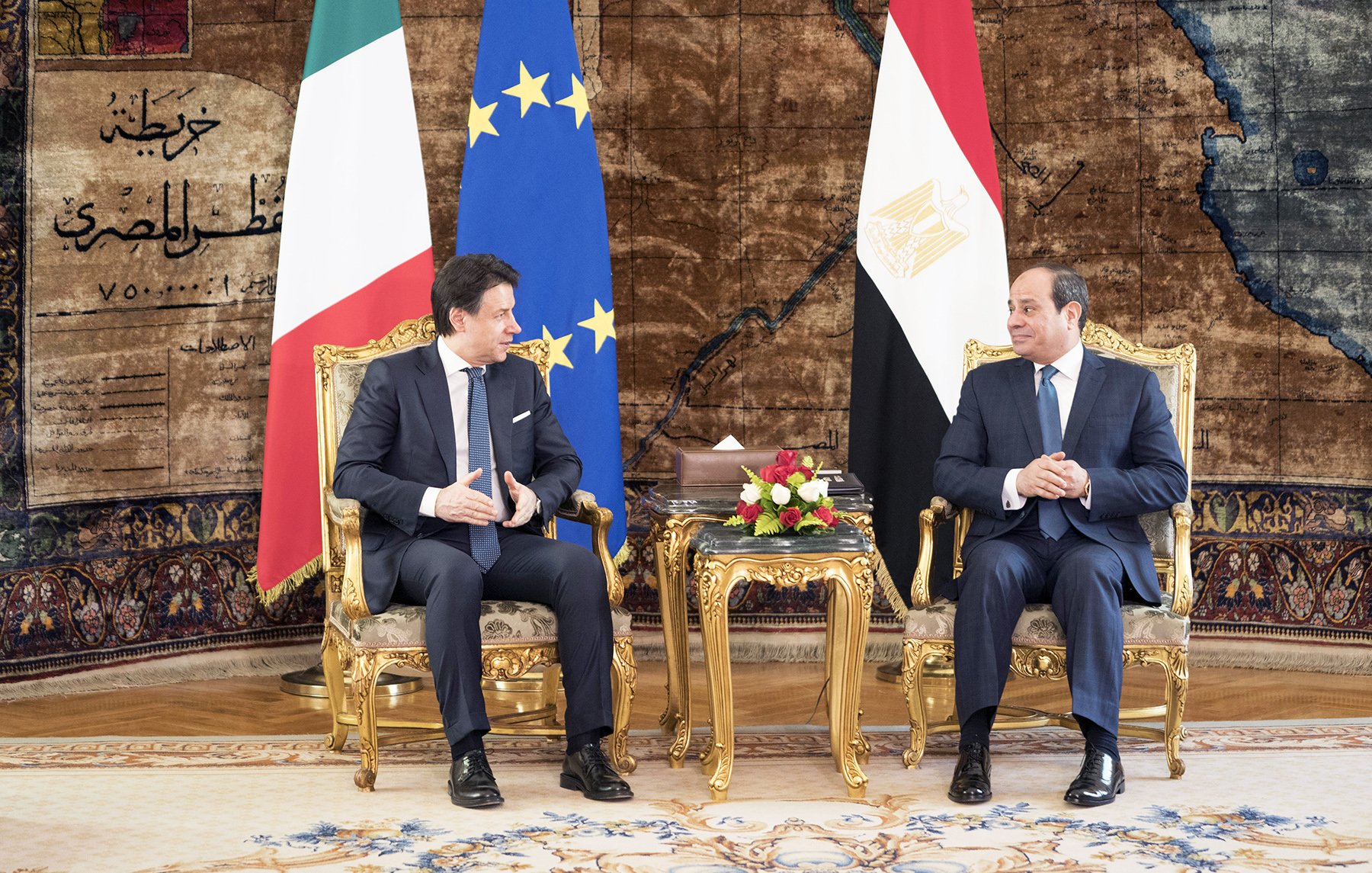 L'immagine mostra due uomini seduti su poltrone dorate in un ambiente elegante. Sullo sfondo si possono notare le bandiere dell'Italia e dell'Egitto, insieme a una bandiera dell'Unione Europea. Entrambi gli uomini stanno conversando e sembrano impegnati in una discussione formale, circondati da elementi decorativi e un tappeto elaborato. Sulle loro ginocchia ci sono bouquet floreali. L'atmosfera è di cordialità e ufficialità.