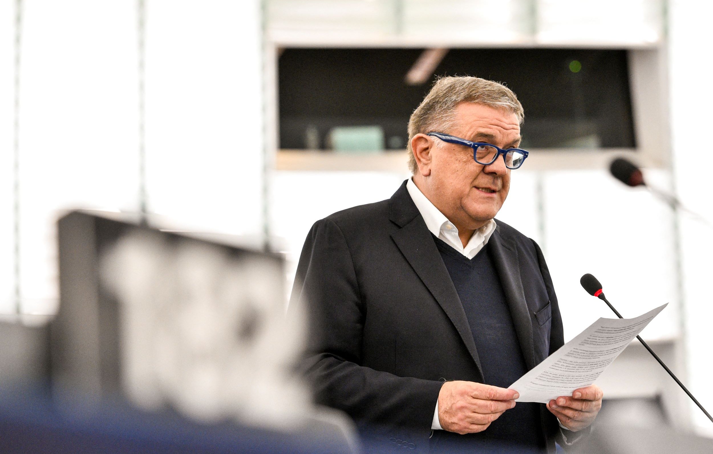 Nell'immagine si vede un uomo che parla a un microfono, probabilmente durante una sessione al Parlamento Europeo. Indossa un blazer scuro e occhiali blu. Tiene in mano un foglio, probabilmente un discorso o una relazione, e sembra essere concentrato mentre si rivolge al pubblico. Sullo sfondo si intravedono alcune strutture dell'ambiente parlamentare.