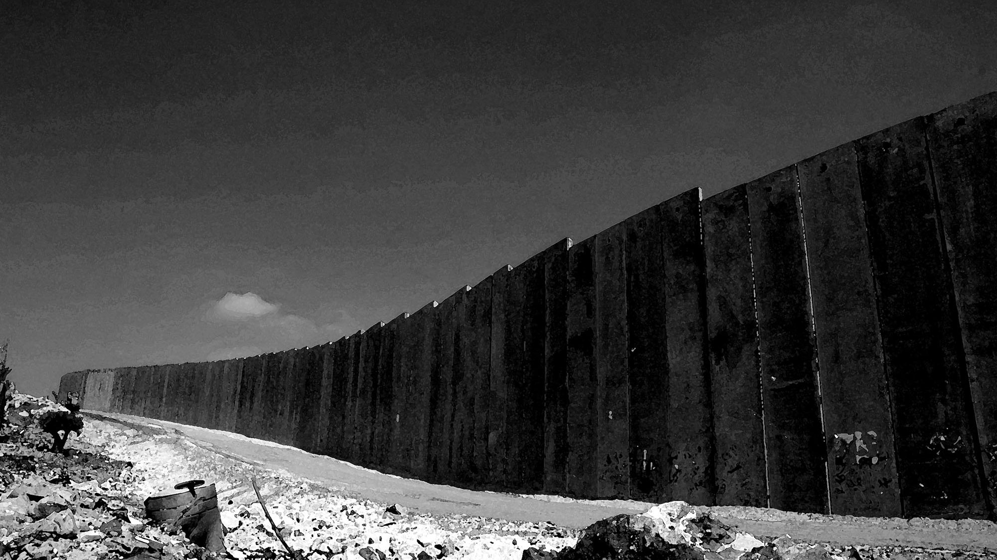 L'immagine mostra un muro lungo e imponente, eretto in un paesaggio desolato. La scena è in bianco e nero, il che le conferisce un'atmosfera drammatica. Il muro, costituito da grandi pannelli di cemento, si estende in modo curvilineo verso l'orizzonte, mentre il terreno attorno appare roccioso e poco curato. In lontananza, si intravede un cielo chiaro, con una nube che si staglia sullo sfondo, aggiungendo profondità all'immagine. L'assenza di colori enfatizza la severità e la rigidità della struttura, suggerendo un senso di divisione e separazione.