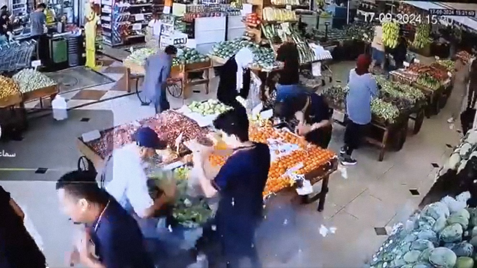 L'immagine mostra l'interno di un negozio di alimentari, probabilmente una fruttivendolo. Ci sono diverse persone che si muovono tra le corsie, mentre vari tipi di frutta e verdura sono esposti su tavoli e scaffali. Le persone sembrano occupate a scegliere e acquistare prodotti freschi. La scena è vivace, con molti colori provenienti dai frutti esposti, come agrumi e verdure di vario tipo. La disposizione degli oggetti suggerisce un'ambientazione ordinata e ben fornita.