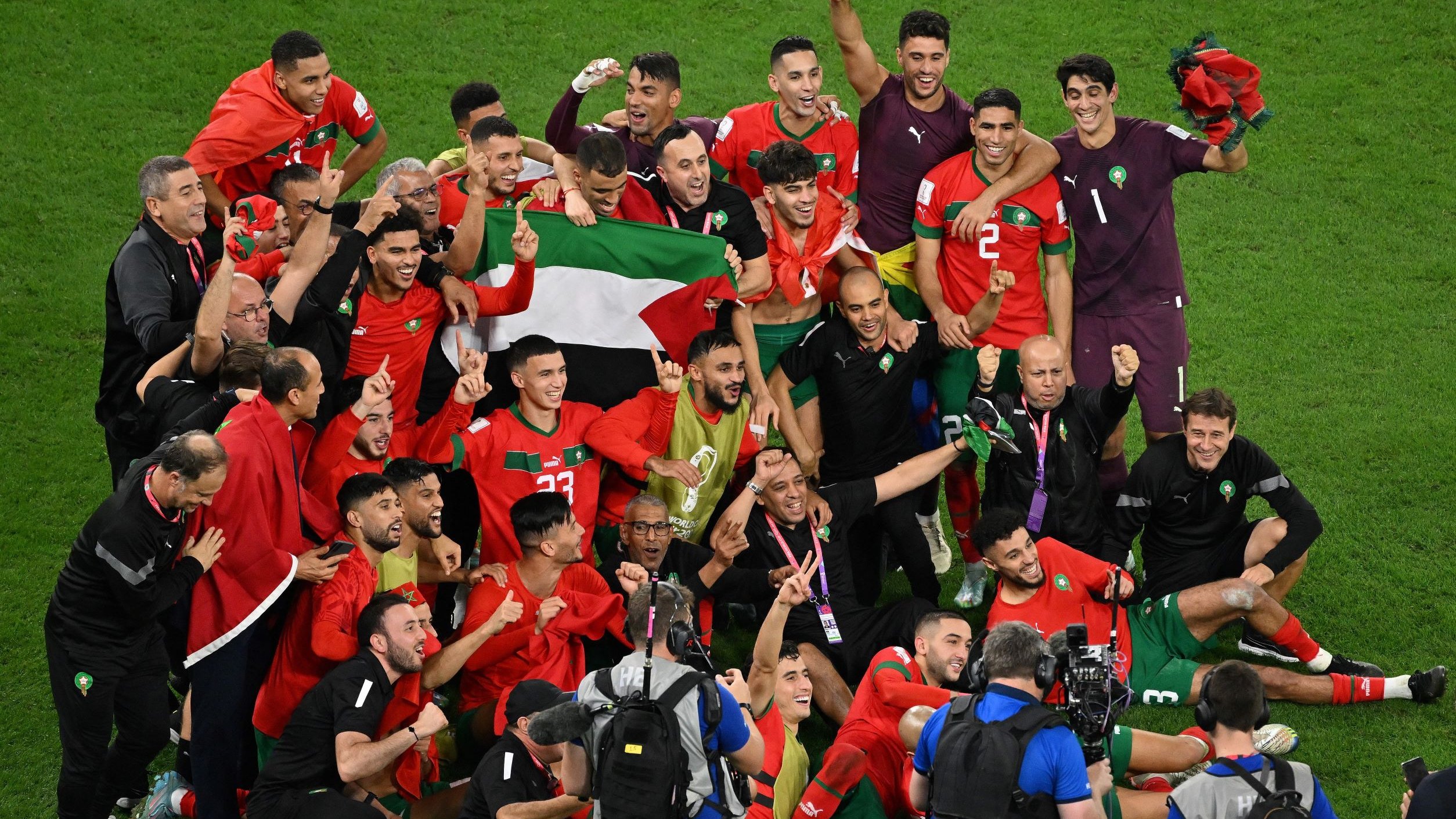 L'immagine mostra un gruppo di giocatori di calcio e membri dello staff, tutti gioiosi e festanti, mentre celebrano una vittoria. Sono vestiti con magliette rosse e verdi, i colori della nazionale marocchina, e alcuni di loro tengono in alto una bandiera. Il gruppo è radunato attorno a un portiere in uniforme, e l'atmosfera è di esultanza e festa. I volti sorridenti e le pose celebrative trasmettono un forte senso di trionfo e unità.