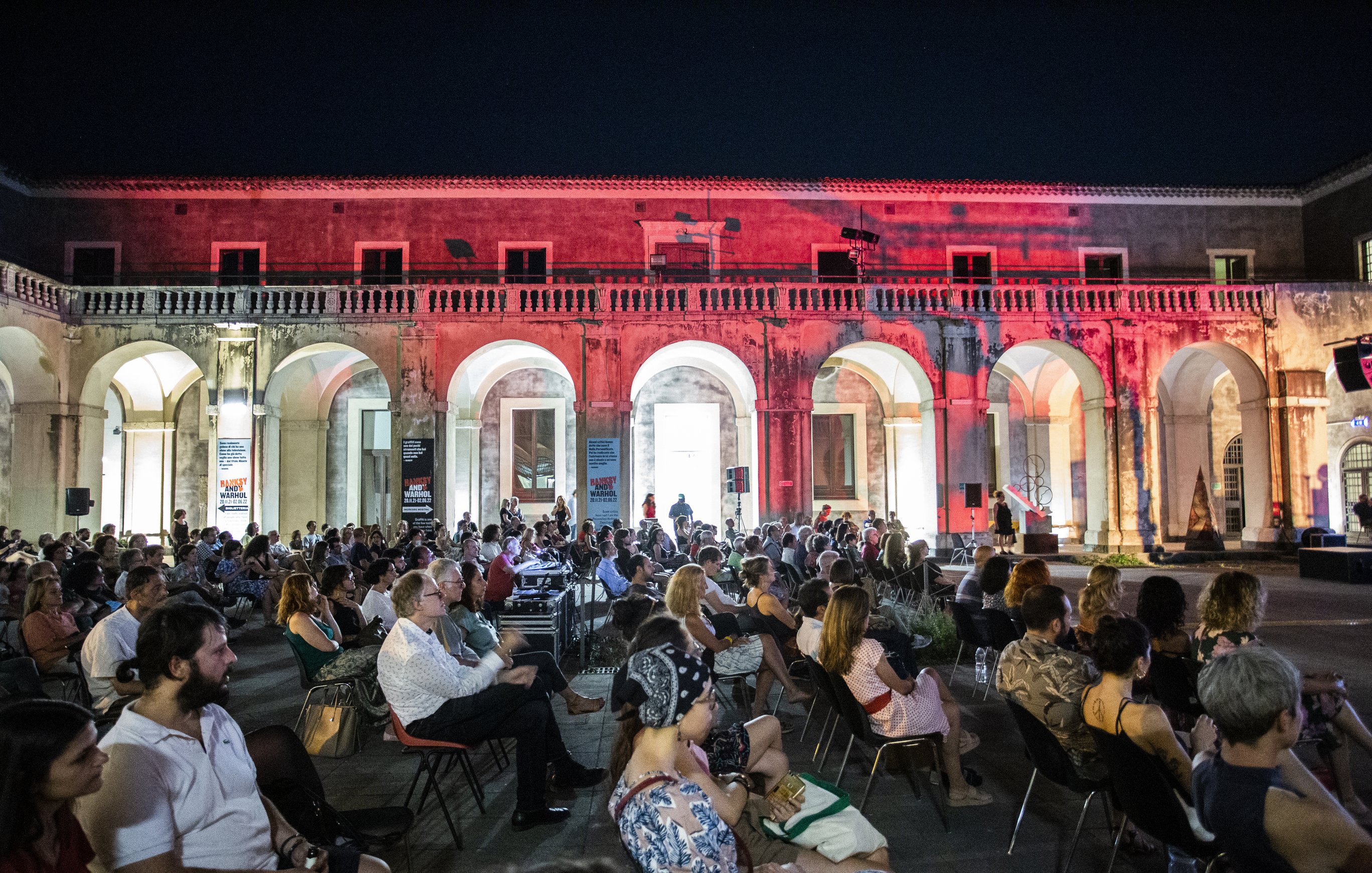 L'immagine mostra una grande sala all'aperto, con archi eleganti che circondano lo spazio. È sera e l'ambiente è illuminato da luci soffuse, creando un'atmosfera suggestiva. In primo piano, un folto pubblico è seduto su sedie disposte in file, mentre alcuni spettatori osservano attentamente. La scena trasmette un senso di partecipazione a un evento culturale o a una performance artistica. I dettagli architettonici dell'ambiente circostante sono ben visibili, contribuendo all'eleganza della location.