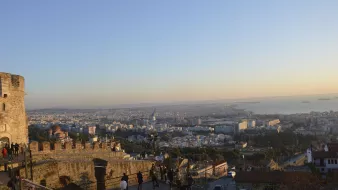 L'immagine mostra una vista panoramica di una città, probabilmente al tramonto. In primo piano si vede una parte di una muraglia o di una fortezza, con alcune persone che passeggiano. Sullo sfondo, si dispiega un vasto panorama urbano con edifici e strade, mentre il mare è visibile all'orizzonte, con alcune navi. I colori caldi del cielo creano un'atmosfera suggestiva, tipica delle ore serali.