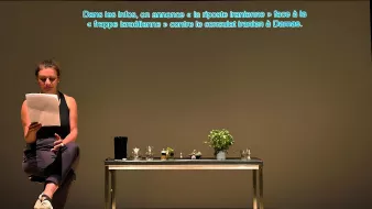 Una donna legge appunti davanti a un tavolo con piante e oggetti, su uno sfondo neutro.