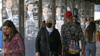 Nell'immagine si vedono diverse persone in un'area urbane, probabilmente un mercato o una strada affollata. Alcuni passanti indossano mascherine, mentre altri non ne hanno. Sullo sfondo, ci sono manifesti con l'immagine di un uomo, presumibilmente un leader politico, appesi alle pareti. L'atmosfera sembra quella di un luogo vivo e multiculturale, con persone di diverse età e stili che si muovono nella zona.