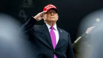 Uomo in abito scuro e cravatta viola, con berretto rosso, fa saluto militare.