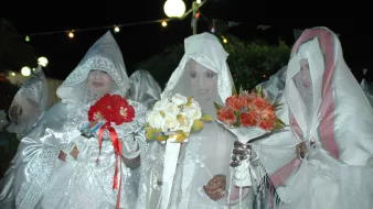 L'immagine mostra un gruppo di donne vestite con abiti bianchi e coperti da veli, che formano un'atmosfera suggestiva. Alcune di loro tengono in mano mazzi di fiori colorati, come rose rosse e fiori bianchi. L'illuminazione è soffusa, creando un'atmosfera notturna, e sullo sfondo si possono vedere luci decorative. Le donne sembrano partecipare a un evento particolare, probabilmente una celebrazione o un rituale tradizionale.