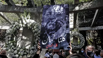 L'immagine mostra una persona che tiene in alto un grande cartello. Il cartello presenta un ritratto in bianco e nero di un uomo con una barba, accompagnato da un testo in arabo che significa "Siamo tutti Libano". Sullo sfondo, ci sono decorazioni floreali e un ambiente che sembra essere un luogo commemorativo. La scena trasmette un senso di unità e solidarietà.