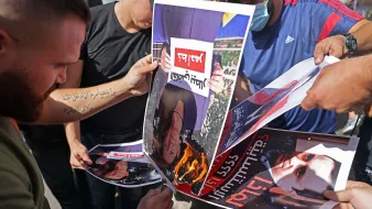 Nell'immagine si vedono diverse persone che stanno bruciando dei manifesti. I manifesti ritraggono volti, probabilmente di una figura pubblica, e sono esposti a una fiamma. Le persone sembrano partecipare attivamente a questa protesta o atto simbolico, esprimendo il loro discontento. L'atmosfera è di intensa emozione e opposizione.