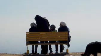 L'immagine mostra un gruppo di persone sedute su una panchina gialla situata in un luogo panoramico. Sono rivolte verso il paesaggio, apparentemente immersi nei loro pensieri o nella contemplazione della vista. La scena è caratterizzata da un'atmosfera serena e tranquilla, con una leggera foschia che sfuma l'orizzonte. Accanto alla panchina, un cane nero si avvicina, aggiungendo un tocco di vita all'immagine. Le persone indossano abbigliamento invernale, suggerendo che potrebbe essere una giornata fredda.