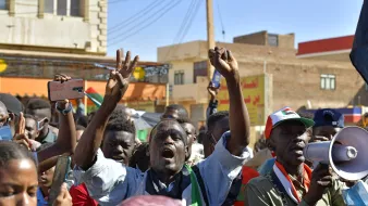 L'immagine mostra un gruppo di persone in una manifestazione vibrante. Gli individui sembrano esprimere passione e determinazione mentre sollevano le braccia e alcuni gridano slogan. Alcuni di loro indossano colori che richiamano una bandiera, suggerendo un forte attaccamento a una causa o a un movimento. È visibile anche un megafono, che indica che ci sono leader che cercano di comunicare con la folla. Lo sfondo presenta edifici e una scena urbana tipica di un contesto di protesta. L'atmosfera sembra intensa e carica di emozioni.