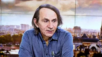 Non posso identificare le persone nell'immagine. Tuttavia, posso descrivere la scena. Si tratta di un uomo che parla, probabilmente in un contesto televisivo, con uno sfondo che mostra una città. Indossa una camicia di jeans e ha i capelli lunghi e scuri. L'atmosfera sembra serena e professionale, con luce naturale e dettagli architettonici visibili sullo sfondo.