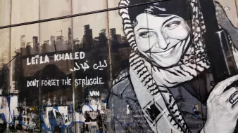 L'immagine mostra un murale che ritrae una donna con un sorriso, indossando un kefiah e tenendo un'arma. Il murale è accompagnato dalla scritta "Leïla Khaled" e dalla frase "Don't forget the struggle", tradotta in arabo. L'opera è situata su una parete, probabilmente in un contesto urbano, e rappresenta un forte simbolo di resistenza e identità. La figura di Leïla Khaled è associata a movimenti di liberazione, in particolare nel contesto palestinese.