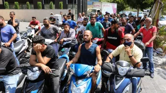 L'immagine mostra un gruppo di persone in fila, molte delle quali sono sedute su motociclette. Sembra che stiano aspettando per ricevere un servizio o un prodotto, probabilmente in un contesto urbano. Le espressioni dei volti variano: alcuni sembrano impazienti, mentre altri appaiono più rilassati. Sullo sfondo si vedono edifici e un'auto, contribuendo a creare un'atmosfera di vita cittadina.