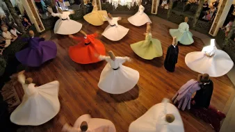 L'immagine mostra un gruppo di ballerini che si esibiscono in una danza tradizionale, con abiti ampi e colorati che girano in cerchio. I ballerini sono disposti in un grande spazio circolare, con un pavimento in legno lucido e una decorazione elegante attorno a loro. Al centro della scena, un lampadario di cristallo illumina l'ambiente, mentre il pubblico osserva l'esibizione con interesse. I colori degli abiti, che vanno dal bianco al viola e arancio, creano un'atmosfera vibrante e festosa.
