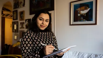 Nell'immagine, una giovane donna sorride mentre tiene un taccuino e una penna in mano. Indossa un maglione con un motivo geometrico. Sullo sfondo si può vedere una stanza con pareti decorate da vari quadri e fotografie. C'è un divano bianco e una sedia gialla, che aggiungono un tocco di colore all'ambiente. L'atmosfera è calda e accogliente.