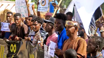 L'immagine ritrae una folla di manifestanti che tiene in mano cartelli e striscioni. I partecipanti sembrano coinvolti ed esprimono entusiasmo, alcuni sorridono mentre altri alzano le mani. Sono visibili diverse espressioni facciali e abbigliamento vario, con colori e stili che riflettono un'atmosfera di protesta. Sullo sfondo si intravede un ambiente urbano, con edifici e vegetazione. L'atteggiamento dei manifestanti suggerisce che stanno lottando per una causa importante.