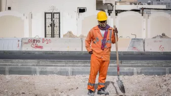 Nell'immagine si vede un lavoratore in un cantiere edile. Indossa una tuta arancione e un casco giallo per la sicurezza. Ha in mano una pala e sembra essere in pausa, osservando il terreno di fronte a lui. Sullo sfondo si possono notare alcune strutture e graffiti su un muro. L'ambiente appare polveroso e in fase di costruzione.
